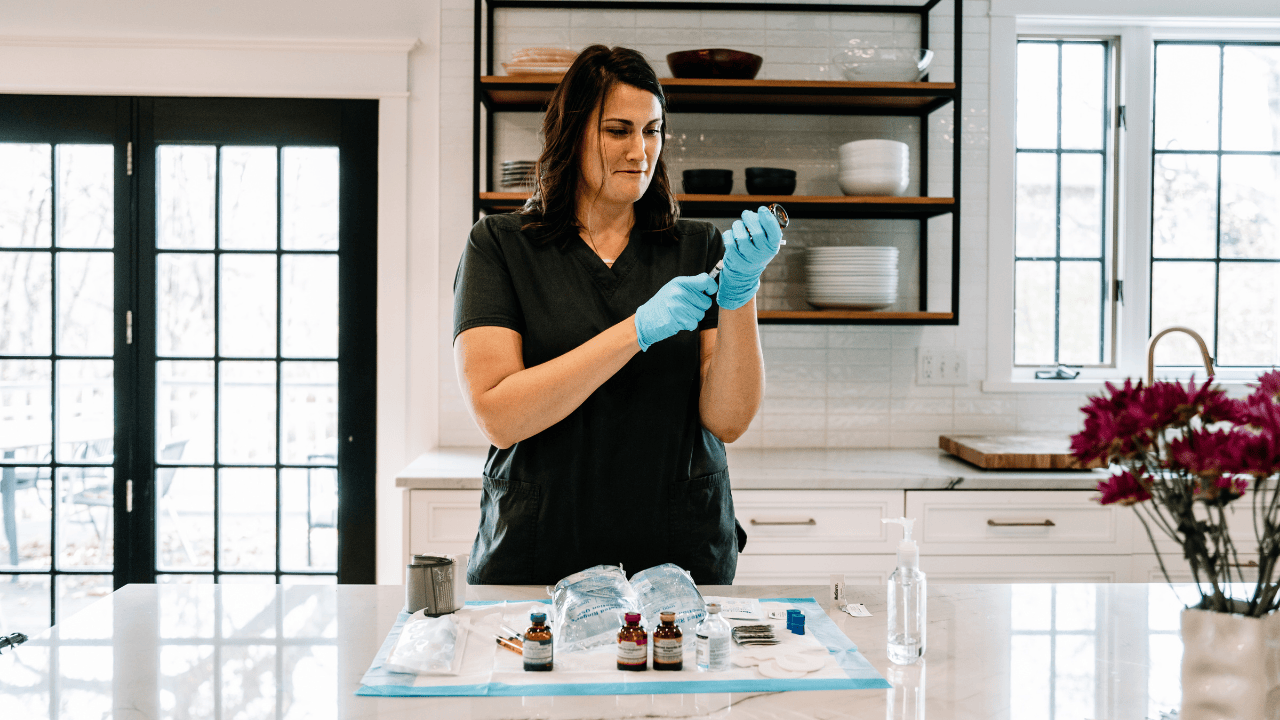 Nurse in black scrubs and blue gloves preparing an IV vial on a kitchen island with medical supplies.